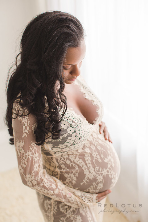 maternity photo of a woman wearing lace dress looking down at her belly neutral colors in a natural light studio by the window
