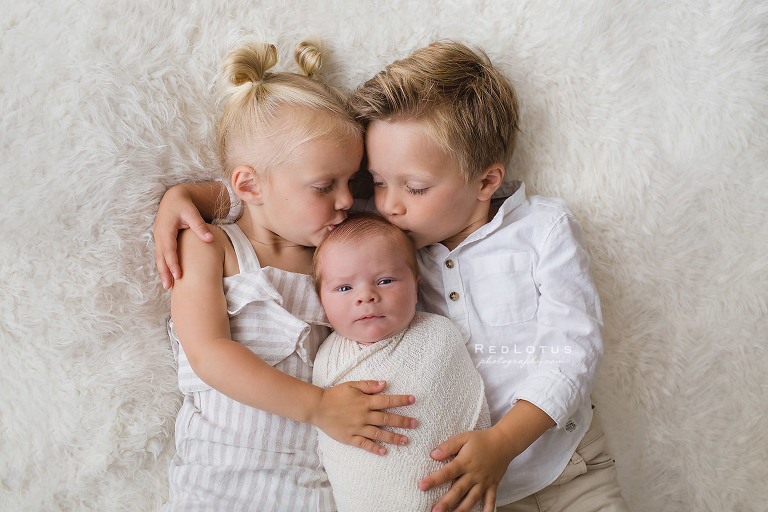 newborn and sibling pose laying down brother and sister kissing baby