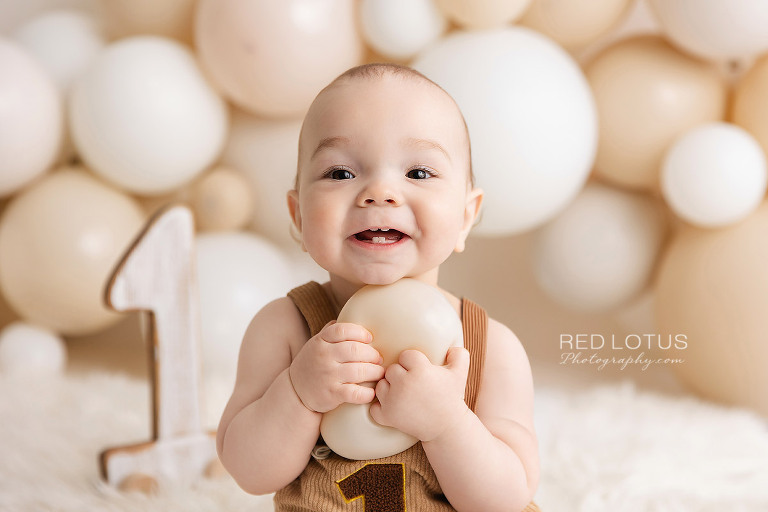 happy one year old baby boy with a neutral balloon garland backdrop