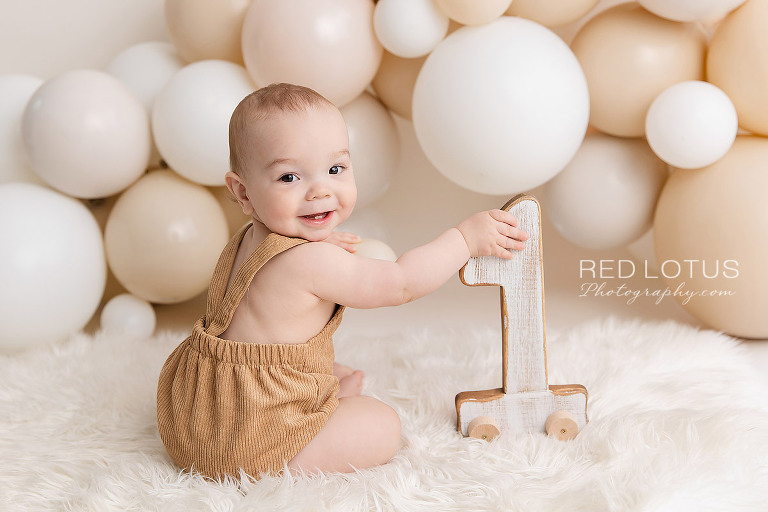 happy one year old baby boy with a neutral balloon garland backdrop