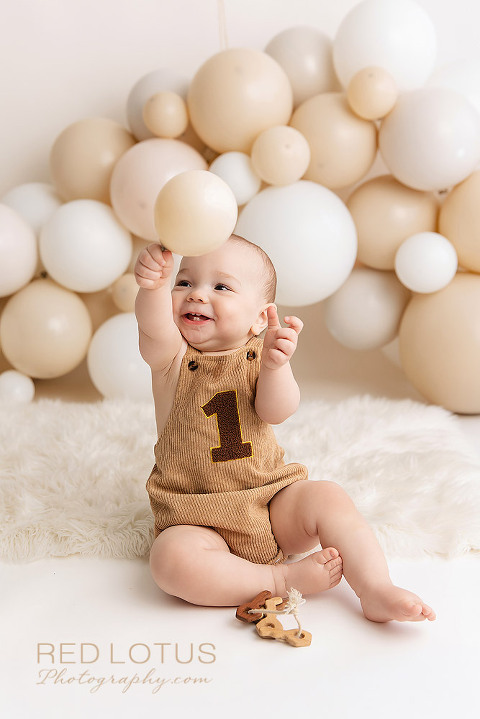 happy one year old baby boy with a neutral balloon garland backdrop