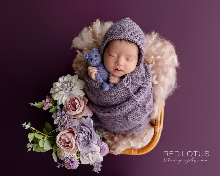 Newborn baby girl in lavender purple swaddle warp with matching bonnet holding teddy bear posed in basket with purple and pink flowers