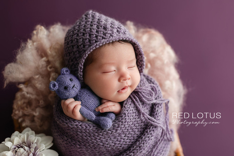 Newborn baby girl in lavender purple swaddle warp with matching bonnet holding teddy bear