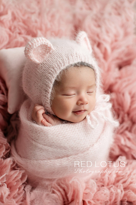 newborn baby girl in pink swaddle wrap with pink teddy bear bonnet smiling
