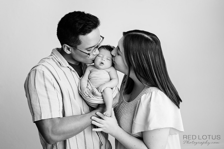 mother and father pose holding baby in the middle kissing baby's cheeks