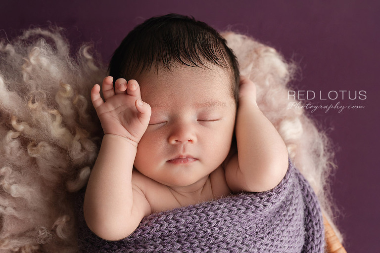 newborn baby girl in a lavender purple swaddle asleep with hand resting on her face