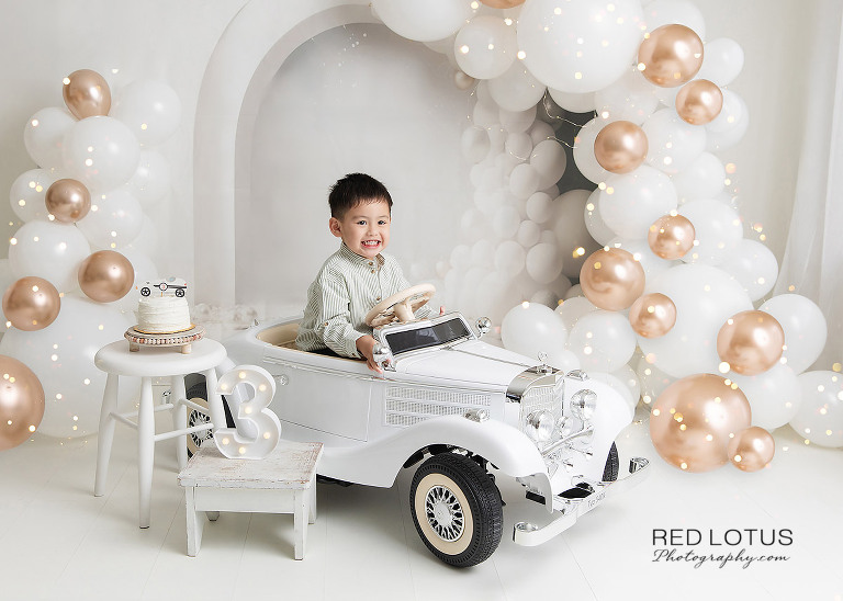 three year old by wearing sunglasses sitting in his toy car celebrating his third birthday with balloons and cake