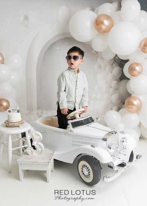 three year old by wearing sunglasses standing in his toy car celebrating his third birthday with balloons and cake