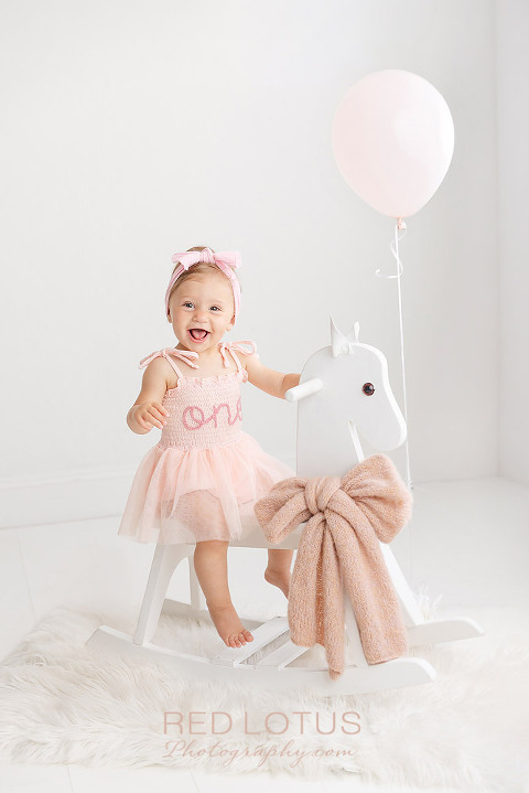 one year old girl in pink dress riding a white rocking horse smiling