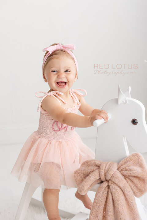 one year old girl in pink dress riding a white rocking horse smiling